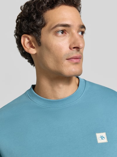 Männermodel trägt Sweatshirt mit Label-Patch von Scotch & Soda in hellem Blau, reine Baumwolle, lockerer Schnitt, Nahaufnahme Oberkörper.