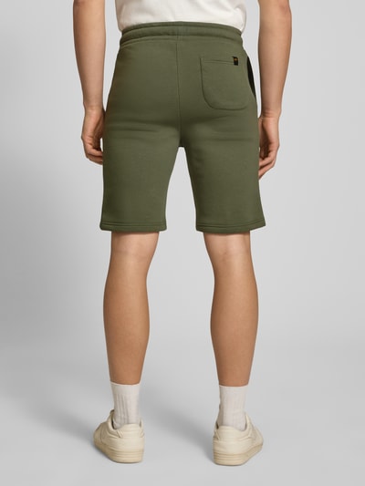 Männermodel trägt Shorts mit Eingrifftaschen und Logo-Print von Alpha Industries in Olivgrün, entspannter Schnitt, Rückansicht Ganzkörperansicht. Baumwoll-Mix, elastischer Bund mit Kordelzug, Gesäßtasche, glatte Textur, Größe M mit 39 cm Bundweite und 24 cm Innenbeinlänge.