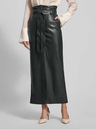 Frauenmodel trägt eng anliegenden Midi-Rock mit seitlichen Eingrifftaschen Modell 'VACARO' von BOSS Orange, Optik: Unifarben, metallische Akzente, Label-Stitching-Applikation, Gehschlitz, Gürtelschlaufen, Bindegürtel, Verschluss: Knopf und verdeckter Reißverschluss, Materialverarbeitung: Kunstleder, dunkle Farbe, glatte Textur, Ganzkörperansicht von vorne.