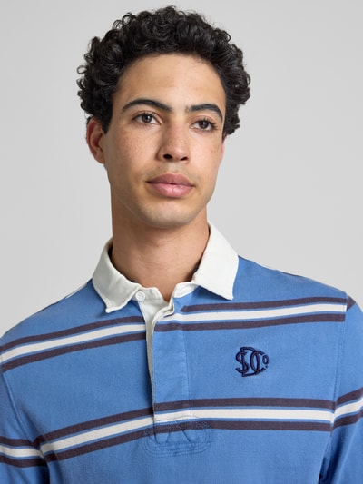 Männermodel trägt Regular Fit Poloshirt mit Logo-Stitching von Superdry in Blau mit weißen und braunen Streifen, reine Baumwolle, Nahaufnahme Oberkörper.