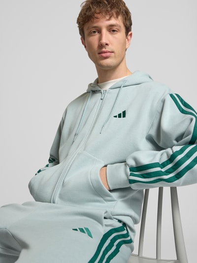 Männermodel trägt Regular Fit Sweatjacke mit Viskose-Anteil von adidas Sportswear, hellblaue Farbe, Logo-Stitching, geteilter Känguru-Tasche, Reißverschluss, elastische Abschlüsse, Baumwoll-Mix mit Viskose-Anteil, Rückenlänge 68 cm, Ärmellänge 60 cm, Nahaufnahme Oberkörper.