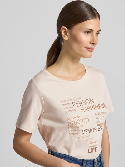 Frauenmodel trägt T-Shirt mit Strasssteinbesatz von monari in Beige mit Statement-Print und Rundhalsausschnitt, Nahaufnahme Oberkörper.