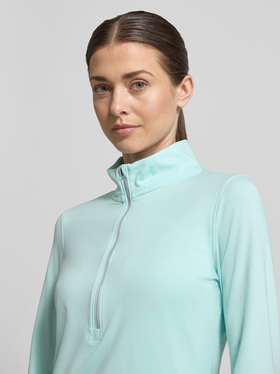 Frauenmodel trägt Longsleeve mit Stehkragen und Reißverschluss von Under Armour in hellem Türkis, Material-Mix mit Stretch, Logo sichtbar, Nahaufnahme Oberkörper.