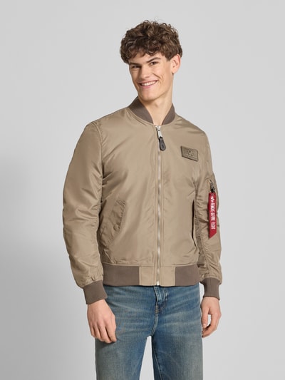 Männermodel trägt Regular Fit Bomberjacke mit Logo-Patch von Alpha Industries in Beige, sichtbarer Reißverschluss, Eingrifftaschen, Ganzkörperansicht von vorne. Rückenlänge 65 cm, Schulterbreite 45 cm, Brustweite 54 cm, Ärmellänge 69 cm.