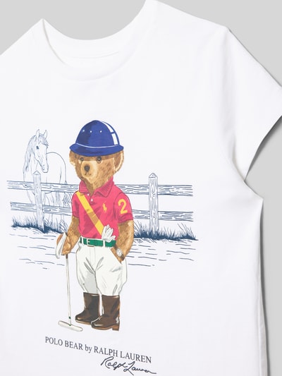Mädchen trägt Dziecięcy t-shirt z nadrukiem z logo Polo Ralph Lauren Teens, bawełna, krój regular fit, okrągły dekolt, całkowita długość tyłu 44 cm przy rozmiarze S, nadruk Polo Bear na tle ogrodzenia, jasne kolory, gładka tekstura. Nahaufnahme Oberkörper.