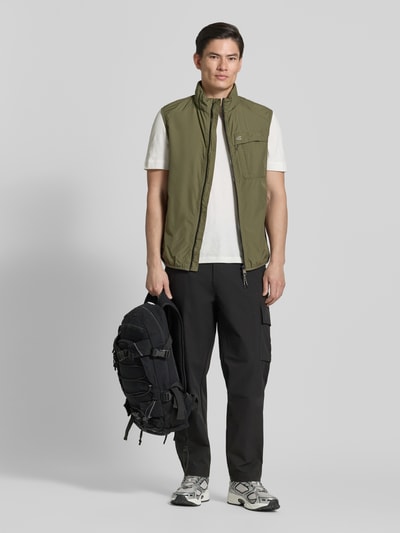Männermodel trägt Regular Fit Weste mit gefüttertem Stehkragen von camel active in Oliv mit Brusttasche, Reißverschluss, Eingrifftaschen, elastischem Bund, Rückenlänge 64 cm, Schulterbreite 45 cm, Brustweite 60 cm; Ganzkörperansicht von vorne.