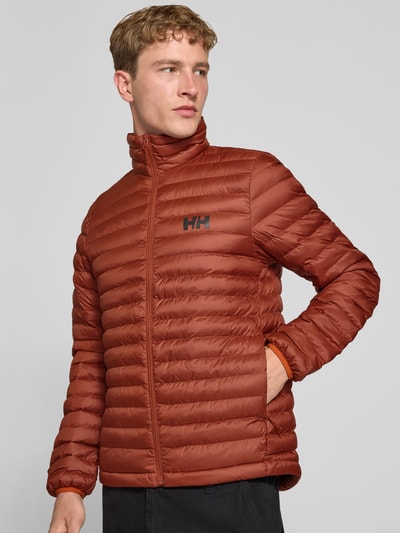 Männermodel trägt Steppjacke mit Label-Print von Helly Hansen in Rostbraun mit Stehkragen und elastischen Ärmelabschlüssen, Ganzkörperansicht von vorne. Gerade Passform, durchgehender Reißverschluss, sichtbarer Label-Print, glatte Struktur, Rückenlänge 72 cm, Schulterbreite 42 cm, Brustweite 54 cm, Ärmellänge 72 cm.