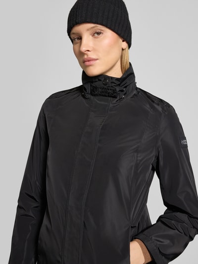 Frauenmodel trägt Regular Fit Mantel mit Logo-Applikation von NAUTGÅRD in Schwarz, Stehkragen, Kapuze mit Tunnelzug, Eingrifftaschen, Nahaufnahme Oberkörper. Rückenlänge in Größe S: 88 cm, Schulterbreite: 37 cm, Brustweite: 48 cm, Ärmellänge: 63 cm.