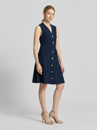 Frauenmodel trägt Knielanges Kleid aus Viskose-Mix Modell 'Amber' von Guess in Dunkelblau mit goldenen Knöpfen, Strick-Optik, ärmellos, taillierte Passform, V-Ausschnitt, knieumspielend, Ganzkörperansicht von vorne.