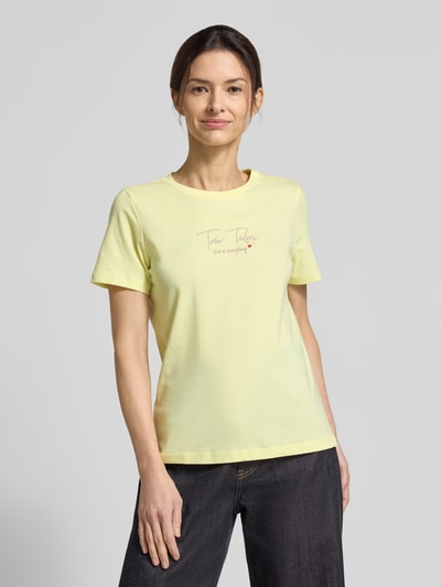 Modelka kobieca nosi Regular Fit T-Shirt aus Bio-Baumwoll-Mix von Tom Tailor, żółty, krój regular fit, krótki rękaw, bawełna organiczna, nadruk z logo, widok całej sylwetki z przodu.