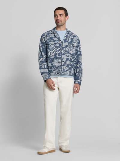 Męski model trägt Jacke im Allover-Look mit Reverskragen von Scotch & Soda, krój regular fit, wzór na całej powierzchni, listwa guzikowa, kieszenie na piersi, długość tyłu w rozmiarze M: 61 cm, szerokość ramion w rozmiarze M: 43 cm, szerokość klatki piersiowej w rozmiarze M: 51 cm, długość rękawów w rozmiarze M: 62 cm, odcienie niebieskiego i bieli, wyraźne wzory i faktura, Ganzkörperansicht von vorne.