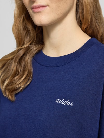 Kobiece modelka nosi Sweatshirt mit Label-Stitching od adidas Sportswear, bawełniany melanż z dodatkiem rayonu, luźny krój, prążkowane wykończenia, okrągły dekolt, subtelne haftowane logo, zbliżenie na górną część ciała.