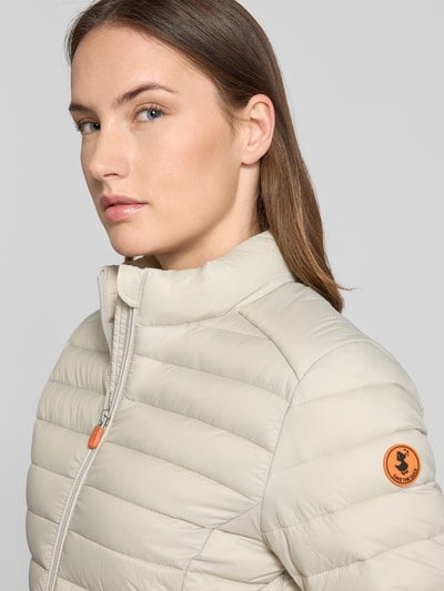 Frauenmodel trägt Slim Fit Steppjacke mit gefüttertem Stehkragen Modell 'Carly' von SAVE THE DUCK in Beige, Stepp-Optik, Nahaufnahme Oberkörper.