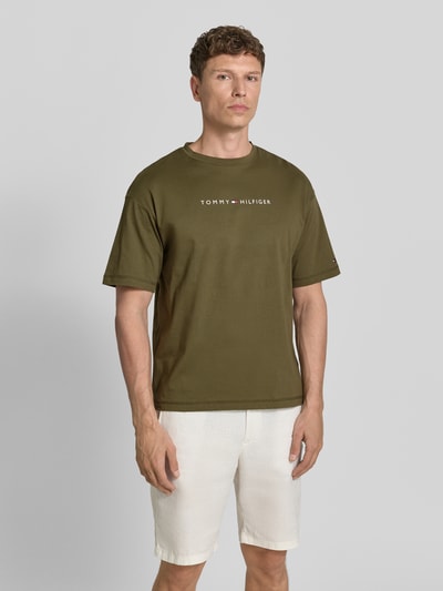 Männermodel trägt Regular Fit T-Shirt aus reiner Baumwolle von Tommy Hilfiger in Olivgrün, unifarben, Label Print, Vorderansicht Ganzkörperansicht.