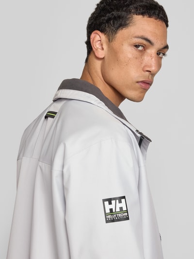 Männermodel trägt Regular Fit Jacke mit Stehkragen Modell 'Crew' von Helly Hansen in Hellgrau, Logo-Print, Nahaufnahme Oberkörper. Herren Jacke von Helly Hansen, Stehkragen, Regular Fit, Modell 'Crew', Logo-Print, Napoleontasche, Eingrifftaschen mit Reißverschluss, Reißverschluss, Rückenlänge bei Größe M: 73 cm, Schulterbreite bei Größe M: 48 cm, Brustweite bei Größe M: 56 cm, Ärmellänge bei Größe M: 68 cm.