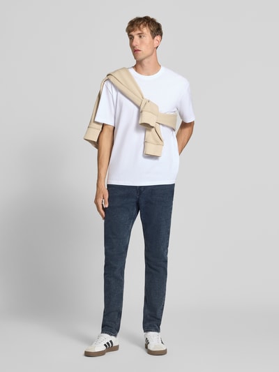 Männermodel trägt Tapered Fit Jeans aus Baumwoll-Mix Modell 'EVEREST' von Calvin Klein Jeans in Dunkelblau mit weißem Shirt, Ganzkörperansicht von vorne, Herren Tapered Fit Jeans von Calvin Klein Jeans.