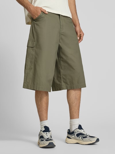 Męski model nosi szorty Straight Fit Shorts mit Cargo-Taschen Modell 'Camo' von KARL KANI z bawełny, z kieszenią cargo, wpuszczanymi i tylną kieszenią, zapięcie na guzik i zamek błyskawiczny, szerokość w rozmiarze M: 41 cm, długość wewnętrzna nogawki w rozmiarze M: 38 cm, kolor khaki, widok całej sylwetki z przodu.