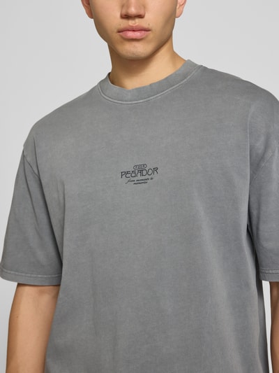 Męski model nosi Oversized T-Shirt mit 1/2-Arm Modell 'Trebic' od Pegador z czystej bawełny, szary kolor, detal z logo, krój oversized, okrągły dekolt, półrękaw, długość tyłu w rozmiarze M: 70 cm, zdjęcie: zbliżenie na górną część ciała z przodu.