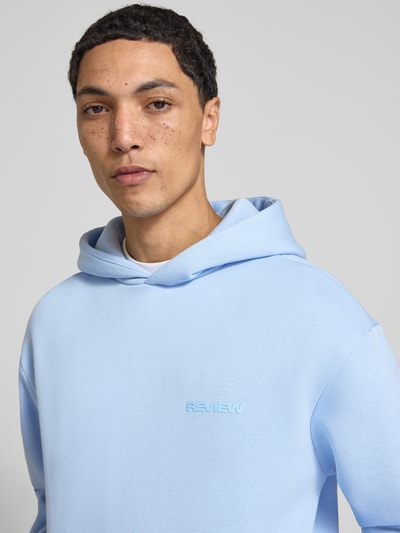 Männermodel trägt Hoodie mit Logo-Print von REVIEW in Hellblau, Baumwoll-Mix, lockere Passform, Nahaufnahme Oberkörper.