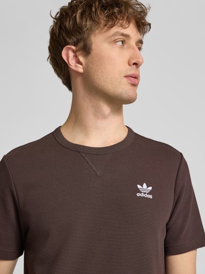 Männermodel trägt Regular Fit T-Shirt mit Baumwoll-Anteil Modell 'AUCO' von adidas Originals, braun, Optik: Unifarben, Struktur, Label-Stitching-Applikation, Rundhals, Kurzarm, Materialverarbeitung: Piqué, Material-Mix; Nahaufnahme Oberkörper.