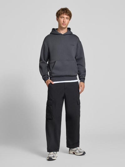Mannenmodel draagt Relaxed Fit Sweatshirt uit katoenmix met kangoeroezak van REVIEW, donkergrijs met labeldetail, vooraanzicht hele lichaam. Ruglengte maat M 68 cm, mouwlengte maat M 62 cm.
