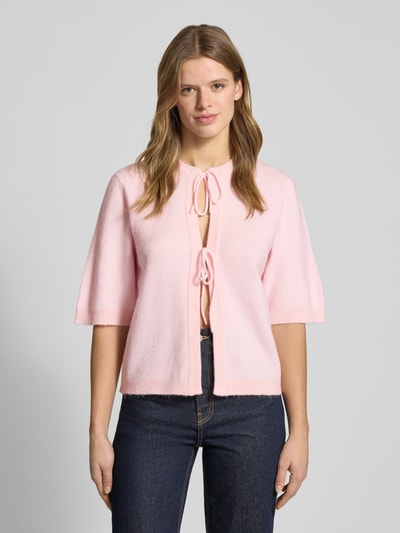 Modelka kobieca nosi Regular Fit Cardigan mit Schnürungen Modell 'SIMONI' od Only, jasnoróżowy, dzianina, krój regular fit, okrągły dekolt, rękaw 1/2, prosta faktura z label stitching, zbliżenie na górną część sylwetki z przodu.
