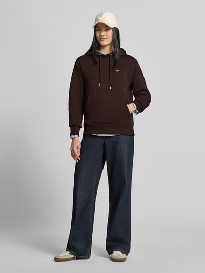Frauenmodel trägt Regular Fit Hoodie aus Baumwoll-Mix von GANT in Dunkelbraun, kombiniert mit blauer weiter Jeans und beigen Accessoires, Ganzkörperansicht von vorne.