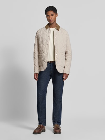 Männermodel trägt Regular Fit Jacke in Stepp-Optik Modell 'Rapid' von CINQUE in Beige mit aufgesetzten Taschen, Ganzkörperansicht von vorne. Elastischer Material-Mix, Umlegekragen und Knopfleiste auf glatter Oberfläche laut Produktbeschreibung.