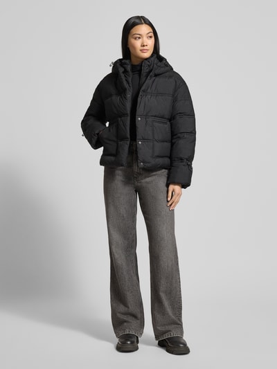 Frauenmodel trägt Cropped Fit Steppjacke mit aufgesetzten Taschen Modell 'Daisy' von Guess in Schwarz mit Allover-Logo-Print, gefütterte Kapuze, Tunnelzug an Kapuze und Ärmelabschlüssen, Ganzkörperansicht von vorne.