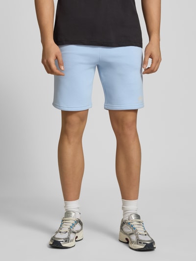 Mannenmodel draagt Shorts mit Eingrifftaschen und Logo-Print van Alpha Industries in lichtblauw katoenmix, relaxed fit, logoprint, steekzakken, elastische band met trekkoord, achterzak, bandwijdte maat M 39 cm, binnenbeenlengte maat M 24 cm. Ganzkörperansicht van voren.