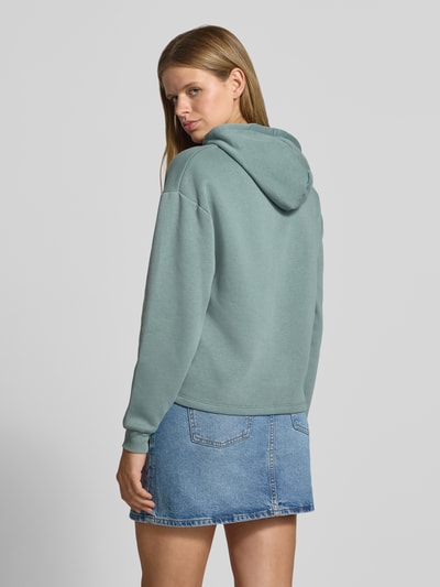 Frauenmodel trägt Regular Fit Hoodie aus Baumwoll-Mix Modell 'CHILLI' von PIECES in hellem Grün zu Jeansrock, Textur weich, Ganzkörperansicht Rückansicht. Damen Hoodie von PIECES.