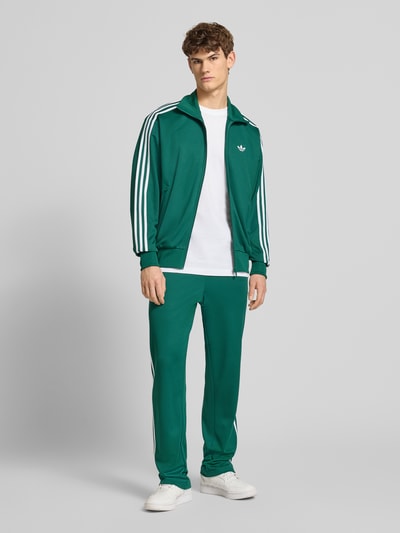 Męski model nosi Trainingsjacke mit Stehkragen od adidas Originals, kolor zielony, krój regular fit, widok całej sylwetki z przodu.