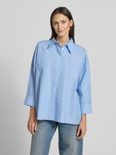 Frauenmodel trägt Relaxed Fit Hemdbluse aus reiner Baumwolle Modell 'Fionne' von OPUS PANTS in Hellblau mit feinen weißen Streifen, Hemdblusenkragen, durchgehender Knopfleiste und 3/4-Arm, ganzkörperansicht von vorne.