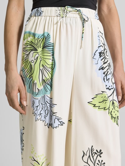 Frauenmodel trägt Stoffhose aus Seide-Mix von HERZENS, cremefarben mit grün-blauem Blumenmuster, elastischer Bund und Tunnelzug, Nahaufnahme Oberkörper. Bundweite bei Größe 36: 37 cm, Innenbeinlänge: 76 cm. Loose Fit Design.