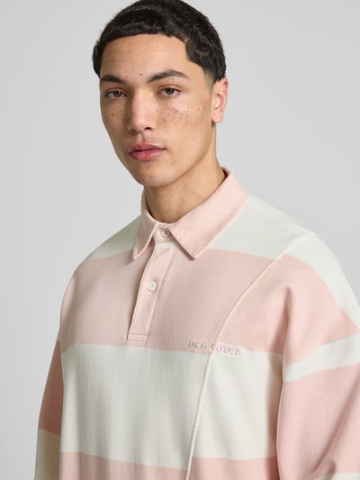 Męski model nosi Relaxed Fit Poloshirt aus reiner Baumwolle Modell 'Castelluccio' od Pegador, pastelowe różowo-białe pasy, kołnierzyk polo, prążkowane mankiety, label-stitching, obniżone ramiona, długość tyłu w rozmiarze M: 69 cm, zbliżenie na górną część ciała.