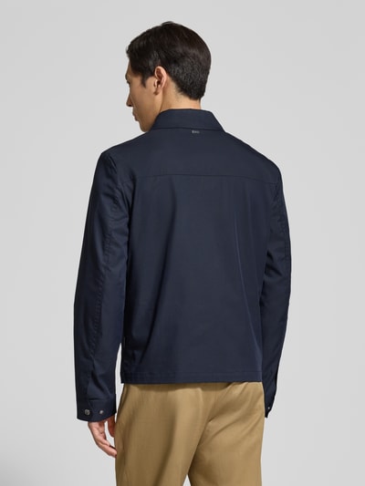 Mężczyznamodel nosi Jacke mit Pattentaschen und Zweiwege-Reißverschluss od bugatti, granatowy bawełniany materiał, prosty krój, patki, zamek dwukierunkowy, długość tyłu w rozmiarze 50: 67 cm, szerokość ramion w rozmiarze 50: 45 cm, szerokość klatki piersiowej w rozmiarze 50: 57 cm, długość rękawa w rozmiarze 50: 70 cm, zdjęcie całej sylwetki z tyłu
