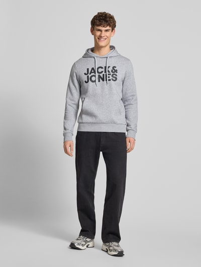 Męski model nosi Hoodie z nadrukiem label i kieszenią kangurką od Jack & Jones, szary kolor, regular fit, widok całej sylwetki z przodu.