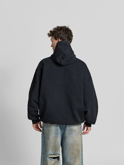 Männermodel trägt Oversized Hoodie aus Baumwoll-Mix von VETEMENTS im Destroyed-Look mit Motiv-Print, dunkle Farben, Rückenlänge 76 cm, Ganzkörperansicht Rückansicht.
