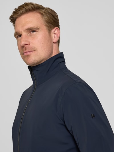 Männermodel trägt dunkelblaue Slim Fit Jacke mit Stehkragen Modell 'Apex' von SCANDINAVIAN EDITION aus Elastischer-Material-Mix, Nahaufnahme Oberkörper, Zweiwege-Reißverschluss, elastische Abschlüsse, Rückenlänge bei Größe M: 65 cm, Schulterbreite bei Größe M: 45 cm, Brustweite bei Größe M: 42 cm, Ärmellänge bei Größe M: 65 cm.