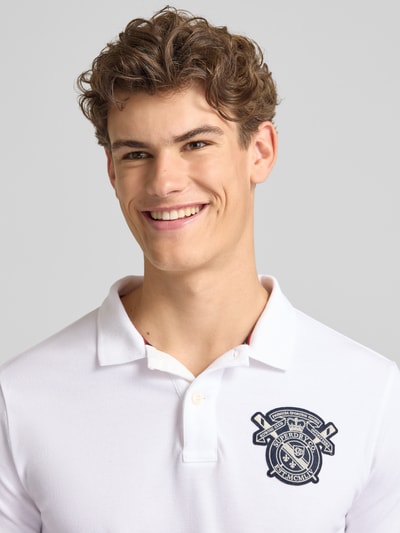 Männermodel trägt Regular Fit Poloshirt mit Label-Patches von Superdry aus Reine Baumwolle, Polokragen, kurze Knopfleiste, Nahaufnahme Oberkörper.