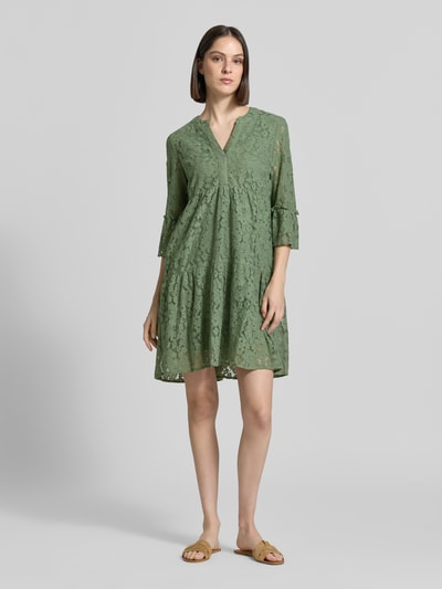 Frauenmodel trägt Loose Fit Spitzenkleid aus Baumwoll-Mix Modell 'HONEY' von Vero Moda in Olivgrün, Spitzenmuster, V-Ausschnitt, Glockenärmel, knielang, Ganzkörperansicht von vorne.