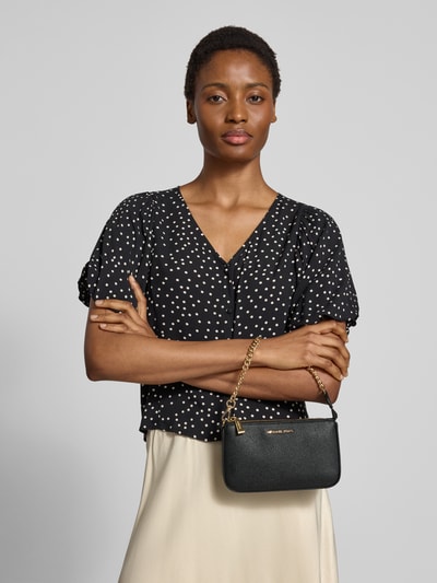 Frauenmodel trägt Shoulder Bag mit Logo-Applikation Modell 'Jet Set' von MICHAEL Michael Kors, schwarze Farbe, goldene Gliederkette, Hauptfach mit Reißverschluss, Maße 20x12x5 cm, Länge Schulterriemen 44 cm, Ganzkörperansicht von vorne.