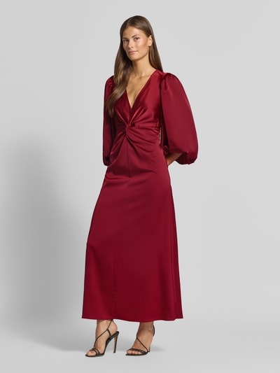 Frauenmodel trägt Cocktailkleid mit Knotendetail und V-Ausschnitt Modell 'THEA' von YAS in Dunkelrot, A-Linie, Maxikleid, Material-Mix mit Stretch-Anteil, leichte Satin-Textur, 3/4-Arm, Ganzkörperansicht von vorne.