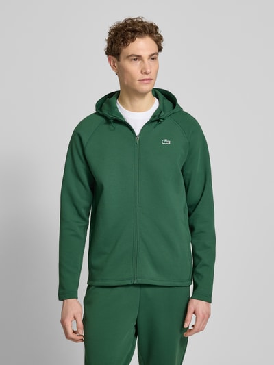 Męski model nosi Regular Fit Sweatjacke mit Baumwoll-Anteil od Lacoste Sport w zielonym kolorze, materiał bawełniany, logo z przodu, pełny zamek, kaptur ze sznurkiem, długie rękawy, boczne kieszenie, krój regular fit, przetwarzanie scuba, widok całej sylwetki z przodu.