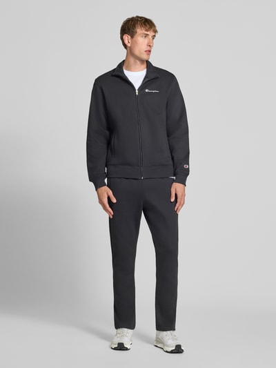 Męski model nosi Sweatanzug mit Label-Details od Champion, kolor czarny, mieszanka bawełny, prosty krój, stójka, zamek błyskawiczny, detal z logo, boczne wpuszczane kieszenie, elastyczny pas, Szerokość w rozmiarze M: 76,42 cm, Długość tyłu w rozmiarze M: 70 cm, Szerokość ramion w rozmiarze M: 51 cm, Długość rękawów w rozmiarze M: 69 cm, Długość wewnętrzna nogawki w rozmiarze M: 76 cm, widok całej sylwetki z przodu.