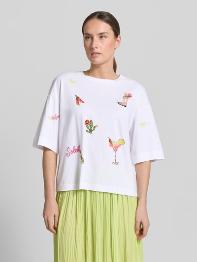 Modelka kobieca ma na sobie T-Shirt mit Motiv-Stitchings od Marc Cain z czystej bawełny, krój loose fit, okrągły dekolt, zdobienia cekinami, biała tkanina, przód zdobiony kolorowymi aplikacjami, długość tyłu w rozmiarze 36: 55 cm, pełny widok z przodu.