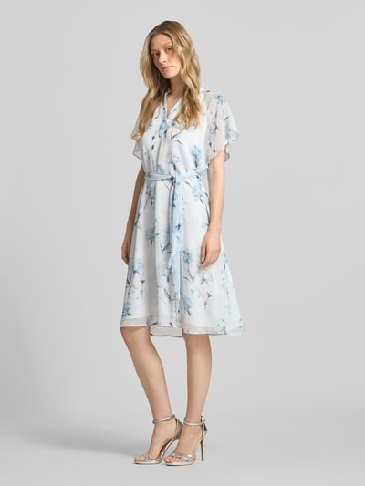Frauenmodel trägt Knielanges Kleid mit Serafino-Kragen von More & More aus leichtem Chiffon mit Schmetterlingsärmel, Taillierter Schnitt, Bindegürtel, helle Blautöne mit Blumenmuster, Knielänge, Ganzkörperansicht von vorne.