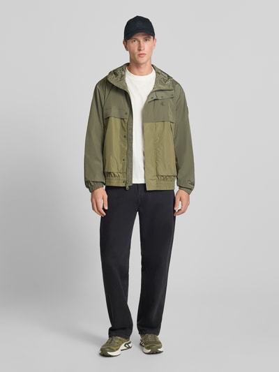 Męski model nosi Regular Fit Regenjacke mit Rückenkoller od Calvin Klein Jeans w odcieniach zieleni, krój regular fit, kaptur z tunelem, jednokolorowy, wzór w kratę, label patch, label stitching, karczek, rękaw długi ze ściągaczem, wykończenie elastyczne, wodoodporna, wiatroszczelna, kieszenie: boczna z zamkiem, wewnętrzna, napoleońska. Ganzkörperansicht von vorne.