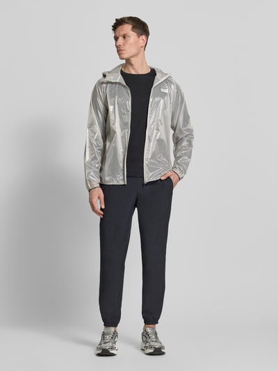 Männermodel trägt semitransparenten Blouson mit Label-Print von EA7 Emporio Armani, Kapuze, Tunnelzug, Reißverschluss, Ganzkörperansicht von vorne.