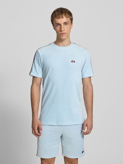 Męski model nosi T-Shirt mit Logo Modell 'MADORI' von Ellesse, mieszanka bawełny z dodatkiem streczu, krój regular fit, okrągły dekolt, logo, długość tyłu w rozmiarze M: 66 cm, jasnoniebieski kolor, prążkowana faktura, widok całej sylwetki od przodu.
